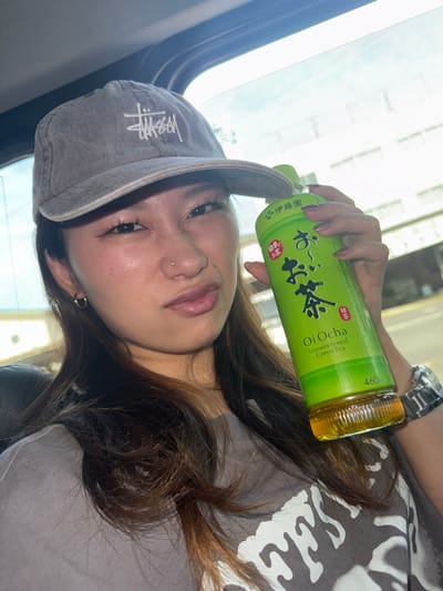 The image shows a young woman wearing a grey "Stussy" baseball cap and a grey t-shirt with white lettering. She is holding a bottle of "Oi Ocha" unsweetened green tea in her right hand. She has dark brown hair and is looking at the camera. The image is taken inside a car with the window open, revealing a view of a street scene outside. The coordinates provided place the scene in Amagasaki, Japan. The woman appears to be in her late teens or early twenties. The car seat is visible in the background.  Her nose has a small stud piercing.  The woman is wearing a gold hoop earring in her left ear.  Her nails are painted light pink.  The woman's expression is neutral.
