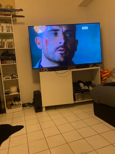 A TV displays a man with a cut on his face. Below the TV is a white shelf filled with various items, including games, consoles, and other electronic equipment. Next to the TV is a white cabinet with an open top. A black speaker is located beneath the cabinet. The room features a tiled floor and a partial view of a sofa in the upper right corner. A black rug is also visible on the floor.