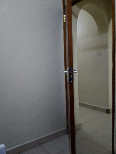 The image displays an empty indoor scene, likely a doorway or entrance to a room within a building. A partially open, dark brown wooden door with a silver-colored handle and visible hinges occupies the right side of the frame. The door jamb is also made of dark wood.

To the left of the door, a plain, light-grey wall fills the majority of the left side of the image. This wall meets a light-colored, possibly off-white, tiled floor at a matching skirting board that runs along the base of the wall. The texture of the wall appears slightly grainy, possibly from the photo quality or the wall finish.

Through the partially open door, another interior space is visible. This area also features light-colored walls and a tiled floor. A prominent feature in the background of this visible space is an arched opening or passage, giving a glimpse into what might be another room or hallway beyond. The lighting in the entire scene is relatively dim and even, suggesting an indoor setting without strong direct sunlight, possibly illuminated by artificial light or indirect ambient light.

There are no people or specific activities depicted, lending a quiet and still atmosphere to the scene. No discernible text is visible anywhere in the image. While the location is contextualized as Kalimoni ward, Kenya, the visual cues within the image itself are generic to many indoor building interiors.