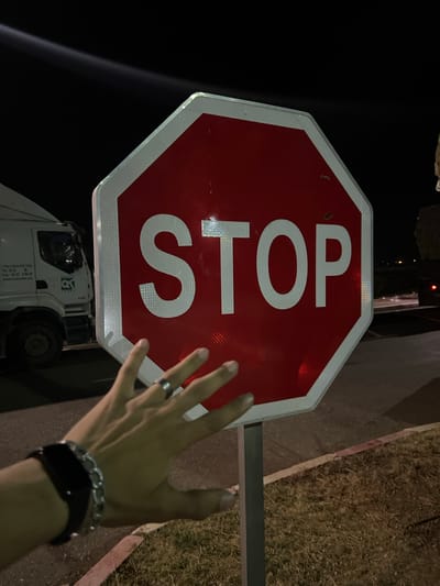 A person extends their hand towards a red and white STOP sign in Jbilate, Morocco. The sign is mounted on a pole. A truck is visible below the sign. The surrounding environment includes pavement, grass, and darkness. The person is wearing a ring, a chain bracelet, and a watch.