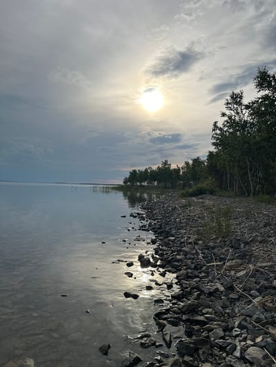 The image shows a natural landscape scene with a body of water and a rocky shoreline. The sun is visible in the sky, casting a bright light and creating reflections on the water. The water appears calm and reflects the sky and surrounding trees. The shoreline is covered with numerous dark gray rocks of various sizes, with some dry grass and thin branches scattered among them. Behind the shoreline, a dense line of green trees forms a backdrop. The sky is partly cloudy, with streaks of white and gray clouds interspersed with patches of blue. The sun's rays are breaking through the clouds, creating a luminous effect. The overall impression is one of a peaceful, natural setting, possibly during sunrise or sunset given the soft lighting. There is no visible text in the image.