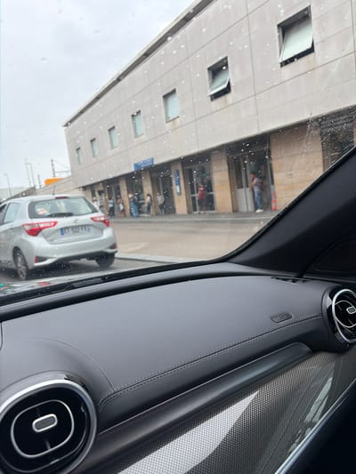 The image shows a view from inside a car, looking out at a street in Villeneuve-Saint-Georges, France. The car's dashboard is visible in the foreground, with a gray textured surface and a circular air vent. Outside the car, a silver hatchback is parked on the street. In the background, a modern building with a beige facade and multiple windows stands. Several people are seen standing in front of the building, some appearing to be waiting. The sky is overcast, and there are raindrops on the car's windshield, suggesting it is raining. The license plate of the silver car is visible and reads "ET 522 FX".