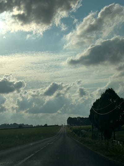 The image shows a view from a car on a road in the countryside, with a drone possibly in the distance or obscured by clouds. The sky is filled with dramatic, grey and white clouds, with patches of lighter sky and sunlight filtering through. A dark green tree, likely a conifer, is visible on the right side of the frame, with power lines strung across it. The road stretches into the distance, leading towards a line of trees and fields. The overall impression is one of a slightly overcast day in a rural setting. The location is stated to be in the city of Grigneuseville, France.