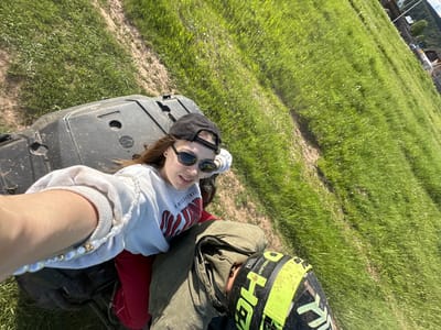 The image depicts two people on an all-terrain vehicle (ATV) in a grassy field. One person is lying on the front of the ATV, wearing a light gray sweatshirt, a black cap, and sunglasses. The other person is sitting behind, wearing a helmet and green outerwear. The location appears to be a rural or semi-rural setting, with green grass covering most of the visible area. A structure is visible in the distance. The image does not provide sufficient information to confirm the location as the city of Пепеляева, Russia.