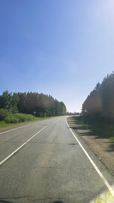 The image depicts a two-lane asphalt road curving to the right, bordered by dense green trees on both sides. The sky above is a clear, pale blue. On the left side of the road, there are signs with red circular markers, indicating a turn. Further down the road, on the right side, there is a triangular warning sign. The road surface shows signs of wear with visible cracks. The scene appears to be in a rural or forested area, possibly on the outskirts of a city. The lighting suggests it is daytime, with the sun casting shadows from the trees onto the road. The specific location in the city of Актюбинский, Russia cannot be determined solely from the visual information in the image.