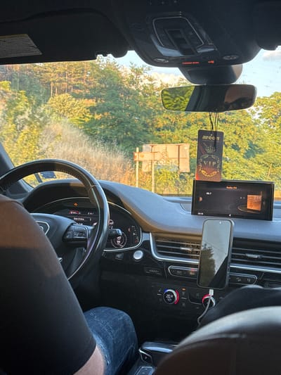 The image presents the interior of an Audi Q7 vehicle, observed from the front passenger seat, situated in Strumyani, Bulgaria. Through the windshield, a dense forest bathed in golden hour light is visible, with a faded brown road sign partially discernible in the mid-ground. The driver is seated, with their left arm in a black t-shirt and legs in blue distressed jeans visible. The steering wheel, featuring the Audi logo, is in the foreground. The instrument cluster displays a range of 240 km, a radio frequency of 88.7, and a speed of 55 km/h, alongside an illuminated warning light (resembling a check engine icon). The central infotainment screen shows "Radio: FM", "88.7", and the time "20:10". An "Areon VIP EXCLUSIVE" air freshener hangs from the rearview mirror. Above the rearview mirror, the overhead console indicates "PASSENGER AIR BAG ON" in red. A smartphone is mounted vertically on the dashboard, actively charging via a white cable. Below, the climate control panel is set to "20.0 AUTO". The windshield exhibits minor specks and dust.