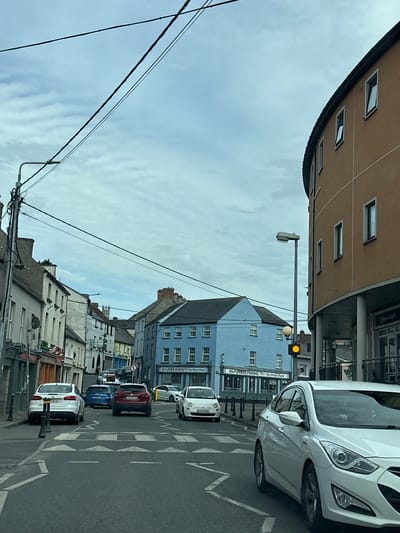 The image depicts a street scene in Carlow, Ireland, likely on a cloudy day. The street is lined with buildings of varying architectural styles and colors. On the right, a modern, curved, ochre-colored building with several windows dominates the foreground. Adjacent to it, a darker-colored building with a shopfront is visible. In the center of the image, a bright blue building with a darker roof and multiple windows stands out. Further down the street, a row of older, narrower buildings in white and cream hues are visible, some with shops at street level.
The street is paved and marked with pedestrian crossings and traffic lines. Several cars are present, including a white hatchback on the left, a red SUV, a blue hatchback, a white sedan in the foreground, and a white Fiat on the far right. A traffic light with a yellow signal illuminated is visible near the blue building. Power lines crisscross the sky above the street. A street lamp is also visible. There are no people clearly visible in the image. The overall impression is a typical European town street with shops and residential buildings.