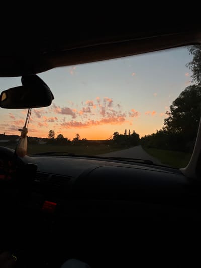 The image is taken from inside a car, looking out through the windshield at a sunset. The sky is filled with wispy clouds tinged with orange and pink from the setting sun. A rural landscape is visible beyond the car's dashboard, with fields, trees, and some houses in the distance. A rear-view mirror hangs from the ceiling of the car, with a small, light-colored object, possibly an air freshener, hanging from it. The interior of the car is dark, with only a red glow visible on the dashboard, suggesting the car's radio or display is on. The time appears to be dusk. The location is identified as Hrabovets, Ukraine.