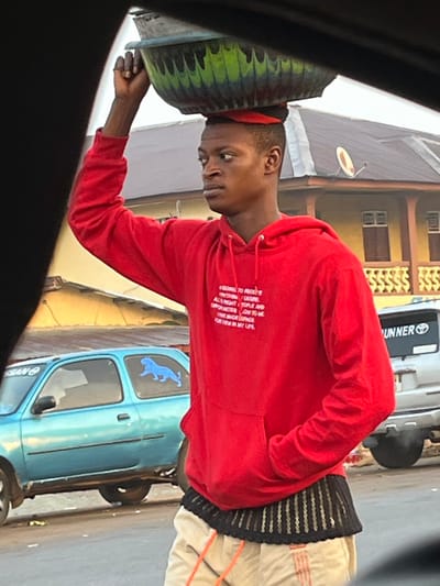 A young man wearing a red hoodie and light-colored pants is carrying a large, round object on his head. He is holding it steady with one hand. The hoodie has writing on the front. Behind him, there is a blue car with a blue dog sticker on the window. A white van with "Runner" printed on the side is also visible. The scene appears to be on a street in an urban setting. Buildings with satellite dishes are in the background. The photo may be taken from inside a vehicle.