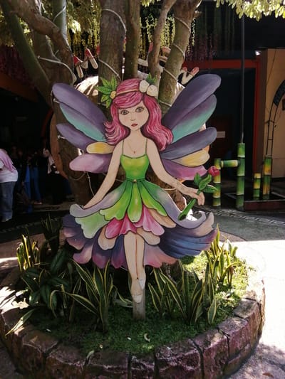 A vibrantly painted, flat cutout of a fairy character stands as a decorative element in a garden setting. The fairy has pink hair, large blue eyes, and wears a green and pink dress that mimics flower petals. Its wings are depicted in shades of purple, blue, and yellow. The fairy is holding a single red rose with green leaves in its right hand. It is positioned in front of a tree trunk that is wrapped with white rope. The fairy cutout is surrounded by a circular stone border filled with green grass and various plants, including snake plants. In the background, people are visible, some wearing headscarves, suggesting a public or semi-public area. There are also decorative bamboo-like structures and building elements in the background. The scene is illuminated by sunlight casting shadows. Based on the provided metadata, this image was taken in Tegalwangi, Indonesia.