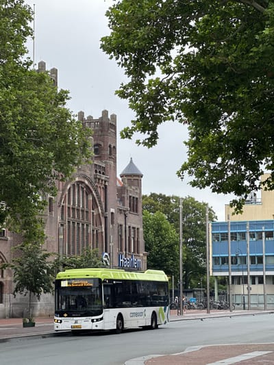 The image shows a street scene in Haarlem, Netherlands, likely near the train station. A modern, white and green electric bus with the Connexxion logo is prominent in the foreground, stopped on the road. The bus displays route information indicating it is route 73 to Castricum via Beverwijk Station. Behind the bus, a distinctive red brick building with Gothic architectural elements and large arched windows stands on the left. The name "Haarlem" is visible on a blue sign mounted on this building. To the right of the brick building, there is a modern blue-glass building. In the background, trees and streetlights are visible, and a row of parked bicycles can be seen near a small bus shelter. The sky is overcast. A person in a light-colored top and blue shorts is standing near the bicycles.