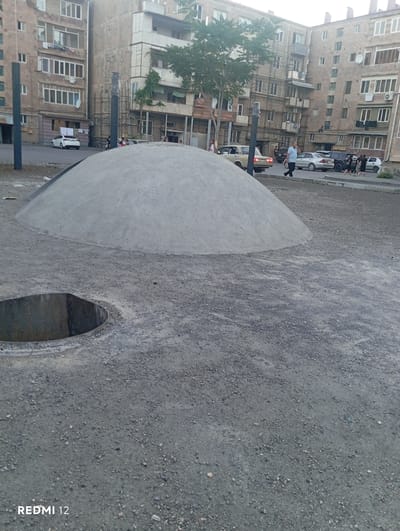 The image depicts an outdoor scene in the city of Vedi, Armenia. In the foreground, a large, dome-shaped mound of gray material, likely sand or concrete, dominates the view. To the left and slightly in front of the mound is a circular opening in the ground, revealing a metal structure. The ground surrounding the mound and the opening is covered in gravel and dirt. In the background, multi-story apartment buildings are visible, characteristic of urban architecture in the region. A paved road runs in front of the buildings, with several cars parked or driving on it. A few individuals are seen walking on the sidewalk or near the buildings. A tree with green foliage is present near the buildings. The lighting suggests it is daytime, possibly late afternoon or early evening. The overall impression is of an urban environment, possibly a construction site or a public space undergoing modification.