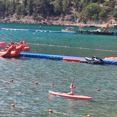 The image depicts a sunny day at a coastal recreation area. In the foreground, a young person is kneeling on a red paddleboard, holding a paddle. The water is calm and appears to be a lake or bay. A rope with red buoys delineates a swimming area. In the mid-ground, a blue floating barrier separates different sections of the water. A black jet ski is docked nearby. To the left, a large inflatable red dragon-shaped water toy is partially visible. In the background, a rocky shoreline is covered with green trees. Further back, a pier with some structures and a small boat are visible. A person is walking on a floating pathway in the far background. The overall scene suggests a leisure activity area at a body of water with various recreational equipment.