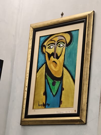 The image shows a framed painting hanging on a textured, off-white wall. The painting is an abstract portrait of a person with a yellow hat, yellow jacket, and blue shirt, rendered in bold, contrasting colors. The subject has a prominent nose and large, expressive eyes. The artwork is signed "haples 1997". A small label with the number "23" is visible in the lower right corner of the matting. The frame is a wide, distressed gold-colored wooden frame. The overall setting appears to be an indoor space, possibly a gallery or home. There is no identifiable context indicating the city of Pozzuoli, Italy.