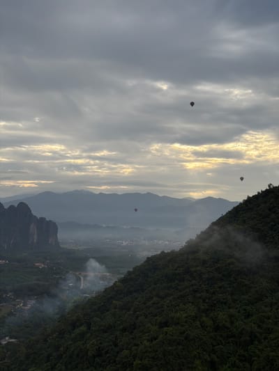 This image captures a panoramic view of Hong Ngern Village, Laos, under a cloudy sky at dawn or dusk. Several hot air balloons are visible in the distance, floating above a misty valley. The foreground is dominated by a dense, dark green forested mountainside, with a lighter green expanse of the village and surrounding terrain in the mid-ground. Layered mountain ranges recede into the hazy distance. Smoke plumes rise from several points in the village, suggesting activity on the ground. The lighting is subdued, with soft, diffused sunlight breaking through the clouds in horizontal bands.