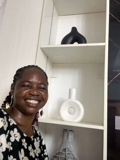 A woman with dark skin and braided hair is smiling. She is wearing a black and white floral print shirt. She is also wearing large gold earrings. Behind her is a white shelf unit containing a black vase, a white vase, and a silver vase. The shelf unit is topped with a black surface with gold marbling. The image was taken indoors. The location is assumed to be Mopo-Akinlade, Nigeria.