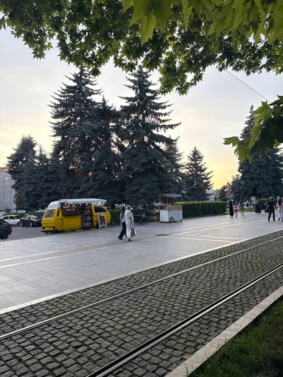 The image depicts an outdoor scene with a street, trees, and several people. The orientation of the image is rotated, with the sky appearing on the left. In the foreground, lush green leaves of a tree partially obscure the view. A paved sidewalk runs alongside a cobblestone street with tram tracks embedded in it.
Several people are visible walking along the sidewalk. A yellow food truck, labeled "SURF BUS COFFEE," is parked on the side of the street, and appears to be open for business. Another smaller cart, possibly a food or drink vendor, is also present further down the sidewalk. The background features a dense line of dark green coniferous trees, and beyond them, buildings can be seen.
The time of day appears to be late afternoon or early evening, with the warm glow of the setting sun visible in the sky, casting a soft light on the scene. The weather seems pleasant, with no indication of rain. The overall atmosphere appears casual and relaxed, with people enjoying an outdoor space. The presence of tram tracks suggests an urban or semi-urban setting.