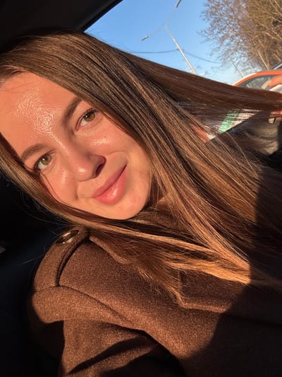 A woman with long, brown hair is pictured from a close-up angle. She has light skin, green eyes, and is wearing a brown coat. Part of a blue sky and some trees are visible in the background. No specific information can be inferred regarding a location in the city of Likino-Dulyovo, Russia.