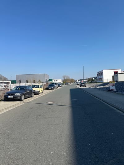 The image displays an asphalt street scene in Schloß Holte-Stukenbrock, Germany, under a clear, bright blue sky containing a faint white aircraft contrail. The street, showing minor surface wear and a manhole cover in the foreground, extends into the distance with a slight upward gradient. A sharp, dark shadow from an unseen structure immediately to the right covers approximately half of the road's width in the foreground.
On the left side of the street, a paved sidewalk borders a series of parked vehicles. These include, from foreground to background, a dark gray BMW 1 Series with license plate GT SE 1812, a yellow two-door hatchback, and several other dark-colored passenger vehicles. Behind these vehicles, a large, light gray rectangular industrial-style building with green accents is visible, followed by a smaller white building.
On the right side of the street, a paved sidewalk transitions to a section featuring a pile of gray gravel and two red and white striped construction barriers in the mid-ground. Further along this side, a white building with red trim along its roofline and multiple windows is visible, followed by another white building in the distance. A tall, dark gray modern streetlight pole stands on the right side of the road in the mid-distance. No individuals are present in the scene.