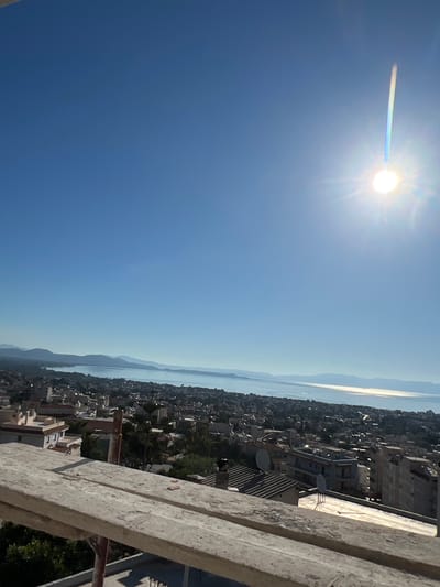 A high-angle, wide shot captures a coastal city under a clear blue sky. The sun, a bright orb with visible rays and lens flare, dominates the upper right quadrant of the image. Below the sun, a faint, vertical rainbow-like streak is visible. The middle ground is a sprawling cityscape with numerous buildings of varying heights and colors, densely packed together. The buildings appear to be a mix of residential and commercial structures, with some exhibiting balconies and satellite dishes. Lush green trees are interspersed throughout the urban landscape. In the background, a serene body of water, likely the sea, stretches to the horizon, bordered by distant, hazy mountains. In the foreground, rough wooden planks of scaffolding are visible, suggesting construction or renovation is underway. This scene is located in Nea Makri Municipal Unit, Greece.