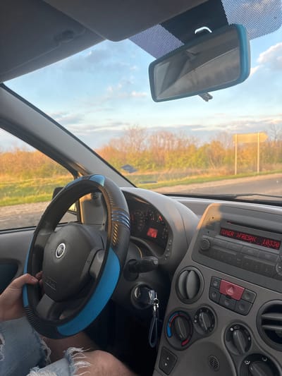 The image shows the interior of a car, presumably in Seltsi, Bulgaria, as suggested by the prompt. The dashboard is visible with a radio displaying "TUNER >FRESH< 10330". Other dashboard components include a hazard light button and climate control knobs. The rearview mirror is in the upper left, and the steering wheel has a blue cover. A person's leg with distressed jeans is visible to the right of the steering wheel. The outside world is visible through the windshield, showing a blurry landscape.
