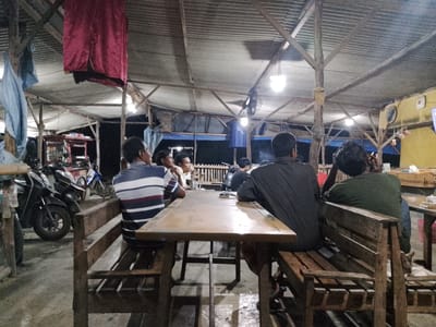 In a dimly lit, open-air establishment with a corrugated metal roof supported by wooden beams, several men are seated at a long wooden table. The setting appears to be a roadside eatery or warung in Sugihwaras, Indonesia. On the left side of the frame, motorcycles are parked. The men are casually dressed, with some facing the table and others looking away. The atmosphere is relaxed, with one man in the foreground on the right wearing a green shirt and facing away from the camera. The floor is concrete and appears wet in places, reflecting the overhead lighting. A small, white cat is visible on a wooden bench to the right of the table. The overall scene depicts a typical evening gathering at a local Indonesian eatery.