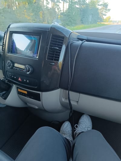 This is a first-person perspective photo taken from inside a vehicle, likely a van or large car. The dashboard is prominent in the foreground, featuring a touchscreen infotainment system, climate controls with dials for A/C and temperature, and various buttons. A black wire is draped over the dashboard.
The driver's feet, clad in light-colored sneakers and grey pants, are visible at the bottom of the frame, resting on a black floor mat. The scene suggests the vehicle is in motion, as indicated by the road and trees visible through the windshield. The sunlight suggests it is daytime, possibly late afternoon given the light quality.
The background shows a road winding through a forested area, with trees on either side. The sky is visible above the trees, appearing bright and possibly with some haze or light clouds. There are no other people or animals clearly visible in the image.
The overall impression is a candid snapshot of a journey, focusing on the interior of the vehicle and the driver's immediate surroundings. There are no strong emotional cues, but the image conveys a sense of travel and everyday movement.
There is some text visible on the infotainment screen, appearing to be a date and time, possibly "20:11 Fri 25.08." Another piece of text is visible on the black cable, reading "MICROPHONE CABLE."