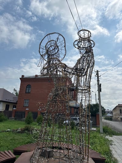 This image shows a large, wireframe sculpture of two figures, possibly a couple, adorned with small white lights. The sculpture is made of metal and appears to be unfinished or designed to be rustic. One figure, on the right, wears a tall hat resembling a top hat, while the figure on the left has a round head covering, possibly a bonnet. They are positioned close together, suggesting an embrace or dance.
The sculpture is situated outdoors in what appears to be a public space. In the background, a red brick building with arched windows stands prominently. To the left of this building, a smaller, lighter-colored house with a sloped roof is visible. Further in the background, a street lined with more buildings, including an older, ornate one on the right, can be seen. Utility poles with wires crisscross the sky above.
The immediate foreground includes a wooden bench constructed from reddish-brown planks. The ground around the sculpture is covered in green grass, with some scattered flowers and small bushes. The sky is partly cloudy, with patches of blue visible through the white clouds. The overall scene suggests a temperate climate, possibly a park or a public square in a town or city. The image was taken from a low angle, looking up at the sculpture and the sky. The location is identified as Nogornyi, Russia.