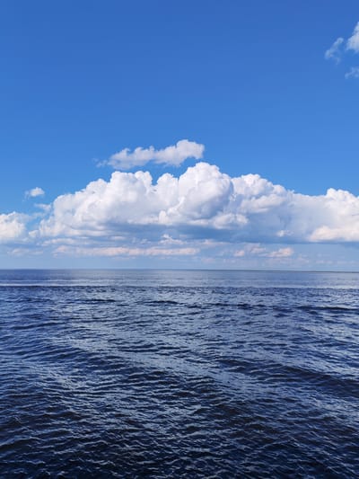 The image depicts a body of water, possibly a lake or sea, under a bright blue sky with scattered cumulus clouds. The water is dark blue with gentle ripples and waves, reflecting the light. A layer of smaller clouds is visible near the horizon. The photo was presumably taken in or near Veregonets, Russia, though no specific location details are discernible in the image. The focus is on the natural scenery. No people or animals are visible.