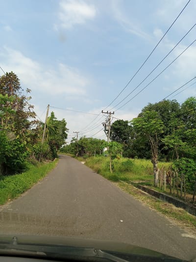 The image depicts a narrow asphalt road in Keudesimpangempat, Indonesia. The road curves slightly ahead and is flanked by dense vegetation on both sides. Utility poles with power lines run along the roadside. The sky is partially cloudy. The view is from inside a vehicle, as evidenced by the dashboard visible at the bottom of the frame. There are no people visible. The environment suggests a rural or semi-rural location.