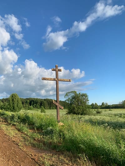 A wooden Orthodox Christian cross stands in a field under a blue sky with white clouds. The cross is dark brown and has a smaller crossbar near the top. Inscribed on the crossbar are golden Cyrillic letters that read "СПАСИ И СОХРАНИ" (Save and Protect). The cross is positioned on a dirt path that curves to the left in the foreground. Behind the cross, a lush green field is filled with tall grass and small white wildflowers, leading to a dense forest in the distance. The scene appears to be in a rural area, likely the village of Svyatitsa, Russia, given the context. No people are visible in the image.