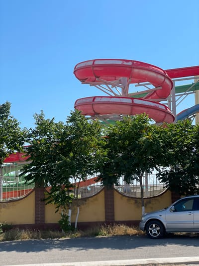 The image shows a sunny day in Ömirzaq, Kazakhstan, with a water slide park visible in the background. Two large, red, curved water slides dominate the upper portion of the frame, with parts of other slides in green and blue visible behind them. The slides appear to be elevated on a metal structure. In the foreground, there is a low, yellow wall topped with a brick pattern, and behind it, lush green trees with full foliage obscure the lower parts of the water slides. A silver SUV is parked on the right side of the image on an asphalt road. The road has a curb and sidewalk area with some dry grass and a few saplings planted along the wall. The sky is clear and blue.