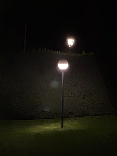 The image depicts a nighttime scene in El Escorial, Spain, featuring two outdoor lampposts against a dark backdrop of a manicured hedge. In the foreground, a tall, slender lamppost with a spherical, frosted glass shade illuminates the green grass below with a soft, yellow light. Further back and slightly higher, a second lamppost with a more angular, lantern-style shade casts a similar warm glow. The hedge forms a dense, dark wall across the midground, its texture visible in the limited light. A solitary wooden post stands vertically to the left of the frame, its purpose unclear. The overall impression is one of quiet outdoor illumination in a residential or park setting.