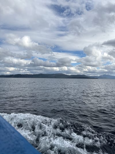 A wide shot captures a body of water with a rocky, mountainous shoreline in the distance. The sky is filled with a mix of white and gray clouds, with patches of blue visible. In the foreground, the blue hull of a boat is visible on the left side, with white wake trailing behind it in the dark water. The scene appears to be from Buvikvoll, Norway.