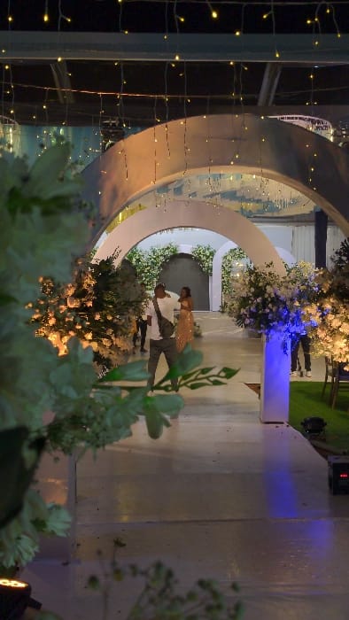 A couple is dancing under an archway decorated with white flowers. The woman is wearing a gold dress, and the man is wearing a suit. The archway is illuminated with lights, and there are more flowers in the foreground. The scene appears to be a wedding or other formal event.