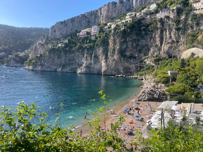 The image displays a panoramic, elevated view of Plage Mala, a beach located in Cap-d'Ail, France. The scene is dominated by a clear, sunlit sky and a crescent-shaped bay with calm, turquoise-to-deep blue waters.

Steep, rocky cliffs covered in sparse vegetation and numerous multi-story residential buildings rise dramatically from the water's edge along the coastline. A small tunnel entrance is visible within the mid-right cliff face.

The beach itself, composed of sand and pebbles, is populated with numerous individuals. Many are engaged in sunbathing on beach towels or sun loungers under a variety of colorful and white umbrellas. Several people are walking along the waterline.

In the bay, numerous individuals are swimming in the clear, shallow waters near the shore. Others are observed on paddleboards or small watercraft. Further out, multiple boats and yachts are anchored in the deeper blue sections of the bay.

Adjacent to the beach on the right, structures associated with a beach club or restaurant are visible, featuring white patio umbrellas, tables, chairs, and dark-roofed buildings. Green foliage partially obscures the immediate foreground, indicating an elevated vantage point for the photograph. The scene is illuminated by bright, natural sunlight, suggesting a clear day.