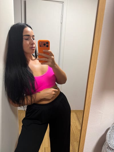 A woman is lying on a white surface, taking a selfie in a mirror. She has long, dark hair, fair skin, and is wearing a bright pink sports bra and black pants. Her left arm is bent, holding an orange smartphone, and her right arm is resting behind her head. A ring is visible on her right ring finger. The reflection in the mirror shows a wooden frame at the top and a white wall behind her. The floor in the reflection appears to be made of wood planks. The lighting suggests it is daytime, likely indoors. The overall mood is casual and relaxed. There is no visible text in the image.