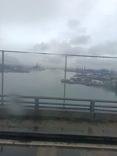 The image depicts an elevated view from a bridge in Arraiján, Panama, looking across a body of water towards extensive industrial port facilities under an overcast sky. In the foreground, the lower section of a concrete bridge structure is visible, topped by a grey metal railing. A chain-link fence with a diamond-shaped mesh pattern extends horizontally across the upper foreground, partially obscuring the view beyond. A blurry artifact, possibly a smudge or water droplet, is present on the lower-left. The midground consists of a calm, grey-blue body of water. Both distant shorelines display significant industrial development. The left shoreline features multiple large gantry cranes at a shipping terminal with several vessels docked. The right shoreline presents a densely developed port area with numerous container cranes, various ships, and associated industrial buildings. The sky is uniformly overcast with grey clouds, indicating diffuse lighting. No individuals or specific human actions are discernible within the frame.