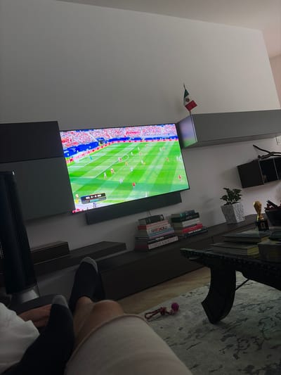 The image depicts a living room scene, likely in Tuxtla Gutiérrez, Mexico, as suggested by the presence of a Mexican flag. In the foreground, two pairs of feet wearing dark socks are visible, suggesting the presence of at least two individuals relaxing. The central focus is a large television screen displaying a soccer match with the score "BEL 0-0 IRN". The television is mounted on a dark gray, cantilevered shelf unit that extends across the wall.
To the right of the television, a small Mexican flag is perched atop the shelf. Below the television, a dark wooden cabinet serves as a surface for several stacks of books, a potted plant in a textured gray pot, and a golden trophy that resembles a miniature World Cup. A dark, ornate coffee table with visible metallic detailing is placed in front of the cabinet and is also covered with books and other items. The floor is covered with a light-colored rug featuring a subtle pattern. To the left of the television, a tall, black cylindrical object, possibly a fan or air purifier, is partially visible. The overall environment suggests a comfortable, modern living space where sports and reading are enjoyed.