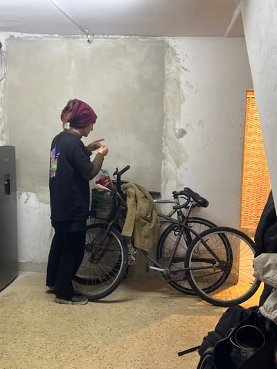 A young woman in a burgundy head covering stands facing the left of the frame, looking at a handheld device. She wears a black long-sleeved shirt over a tan sweater and dark pants. Behind her, against a white, unfinished wall, are two bicycles. One bicycle has a brown jacket draped over it. The environment appears to be an indoor storage area or garage. The location cannot be confirmed to be Victoria, Argentina from the provided image.