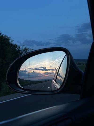 The image depicts a twilight scene captured from the interior of a vehicle, situated in the vicinity of Drugan, Bulgaria. The central focus is the car's passenger-side exterior mirror, which reflects the road and sky behind the vehicle. In the mirror's reflection, a two-lane asphalt road with visible white lane markings recedes into the distance under a vast sky. The reflected sky transitions from deep blue overhead to softer hues of orange, pink, and light blue near the horizon, indicative of a sunset or dusk. Multiple distinct white contrails crisscross the reflected sky. Beyond the mirror, the direct view shows a dark blue twilight sky with scattered, darker clouds. Dark green trees and foliage are visible on the left side of the frame, silhouetted against the sky. The immediate foreground and right side of the image display parts of the car's dark interior, including the door panel and window frame. No individuals are present in the scene. The overall lighting is low, consistent with the late evening hour.