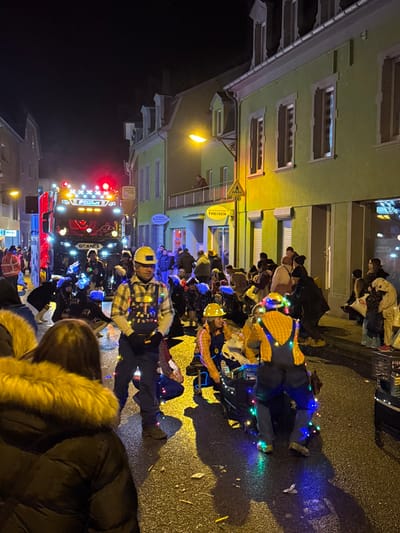 This image depicts a nighttime street scene in Wasserbillig, Luxembourg, characterized by a lively public gathering, likely a parade or festival. The wet asphalt street, reflecting artificial lights, runs through a built-up area lined with multi-story buildings. On the right, a prominent light-green building features several windows with white frames and a balcony on the first floor where a single individual observes the scene. Below the balcony, a yellow sign reads "THEISEN."

In the mid-ground, a group of individuals forms a procession element. A male figure stands centrally, facing slightly right, wearing a yellow hard hat, a plaid shirt, dark overalls, and adorned with multi-colored string lights. To his right and slightly behind him, two other individuals, also in yellow hard hats and overalls, are crouched near a low-lying object or small float that is heavily decorated with blue and multi-colored string lights. Other costumed participants wearing similar attire and illuminated accessories are visible further back.

In the background, a large, dark Scania truck with numerous bright red and white lights on its front and roof leads or is part of the procession. A dense crowd of spectators lines the street on both sides, with many wearing dark winter clothing. The immediate foreground shows the back of a person wearing a dark puffer jacket with a fur-lined hood, looking towards the event. Overhead, streetlights cast a warm yellow glow, illuminating the immediate area. Debris, possibly confetti or discarded items, is scattered on the wet street surface.