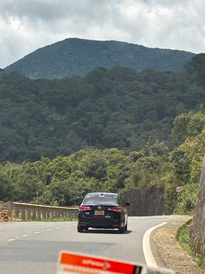The image shows a black sedan driving on a winding road in a mountainous, forested area. The car is positioned in the center of the frame, heading away from the viewer. The road has yellow lane markings and a concrete barrier on the left side. On the right side, there is a stone wall and vegetation. In the background, a large, densely forested mountain rises under a cloudy sky. There is a traffic sign on the right side of the road. The scene appears to be in or near the city of Lac Duong, Vietnam, a region known for its highland scenery and coffee plantations. The overall impression is one of travel through a natural, scenic landscape.