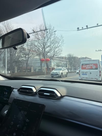 The image provides a forward-facing view from inside a vehicle traveling or stopped on a city street in Huanggu District, Shenyang, Liaoning Province, China.

In the foreground, the vehicle's dark grey dashboard is visible, featuring two prominent hexagonal air vents with silver trim. To the left, a digital display screen shows "5 16," and a larger vertical touchscreen tablet on the lower dashboard displays "13:10" with various application icons. A black rear-view mirror is mounted centrally on the windshield.

Beyond the windshield, a multi-lane asphalt road extends into the distance under a clear, light blue sky. Directly ahead, a white cargo van with a red and white advertisement on its rear and license plate "辽 A 6M258" is visible in the same direction of travel. In the opposing lane, a white SUV is approaching the camera. On the left side of the road, a utility pole supports a black street light. Two red, spherical Chinese lanterns are suspended near this pole. Bare deciduous trees line both sides of the street, and multi-story urban buildings are visible in the background. Overhead, a gantry structure with traffic monitoring equipment spans the road. No individuals are explicitly visible.