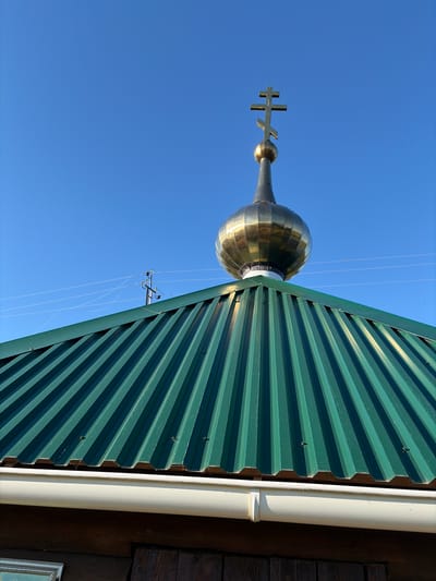 The image shows the top of a church in Pobedinka, Russia. A golden Orthodox cross sits atop a golden dome, which is mounted on a conical spire. The dome and spire are reflective, showing the blue sky and the green metal roof of the church. The roof is corrugated and slopes downwards, with a white gutter running along the edge. In the background, a utility pole with wires can be seen against the clear blue sky. The lower portion of the image shows a wooden wall and a framed picture.