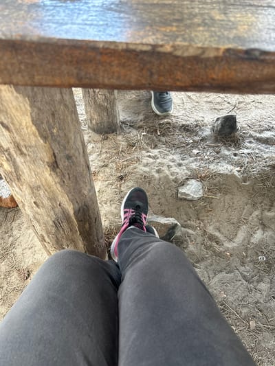 The image shows a first-person perspective of a person sitting on a wooden structure, possibly a bench or picnic table. The person is wearing dark gray sweatpants and black athletic shoes with pink accents. Their legs and feet are visible in the foreground. The ground beneath is sandy and covered with pine needles and small stones. In the background, another person's feet wearing dark sneakers are visible. There are also two large, rough-hewn logs, one to the left and one behind the person in the foreground, which appear to be part of the structure or the surrounding environment. A rough wooden plank runs horizontally across the top of the image, obscuring the upper part of the scene. The overall environment suggests an outdoor, possibly recreational area. The location is identified as Chmyrivka, Ukraine.