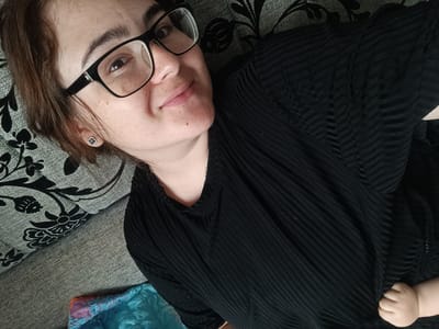 A woman with brown hair and glasses lies on a couch, looking at the camera. She is wearing a black, pleated top and a small, four-leaf clover earring. A baby's hand is visible in the lower right corner of the image. The couch has a grey textured fabric on the left and a black floral pattern on the right. A colorful blanket is partially visible in the lower left. The scene appears to be indoors, in a residential setting. The location in the city of Усть-Тунтор, Russia, is not discernible from the visual information provided in the image.