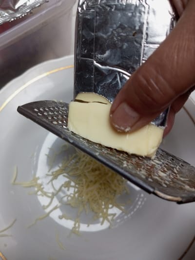 A close-up, high-angle shot captures a culinary activity in the city of Naya andura, India. A human hand is actively grating a rectangular block of light yellow cheese, partially wrapped in silver foil, against a flat, metallic grater. The grater features small, circular perforations. A quantity of finely shredded, light yellow cheese has accumulated on a white ceramic plate, which features a gold rim, positioned directly beneath the grater. A blurred, translucent plastic container with a metallic top edge is partially visible in the upper left quadrant of the frame.