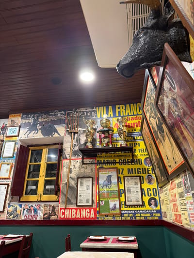 The image depicts the interior of a restaurant or establishment in Santarém, Portugal, heavily themed with Portuguese bullfighting (tauromachy) memorabilia. The location is confirmed by multiple posters bearing the name "SANTARÉM."
The ceiling is constructed from dark, horizontally-laid wooden planks, featuring a circular recessed light fixture and an air conditioning vent. In the upper right corner, a dark, textured bull's head mount with horns is affixed to the wall, partially obscured by framed items.
The walls are almost entirely covered from ceiling to a dark green wainscoting with various posters, framed pictures, and documents, predominantly related to bullfighting events and figures. Notable text includes "VILA FRANCA," "NAZARÉ," "CHIBANGA," "MONTEMOR," and "João Salgueiro," alongside images of cavaleiros (horseback bullfighters), bulls, and forcados.
Centrally, a two-paned window with a bright yellow frame and dark red shutters is visible, with the shutters partially closed. To its right, a rustic wooden pitchfork leans against the wall. Above this, a dark shelf displays three prominent golden trophies of varying sizes, flanked by smaller trophies.
The lower portion of the walls is clad in dark green wainscoting. In the foreground, two restaurant tables are visible, each draped with a white tablecloth and set with a white plate, napkin, and cutlery. Dark reddish-brown wooden chairs are positioned around the tables. No people are present in the scene.