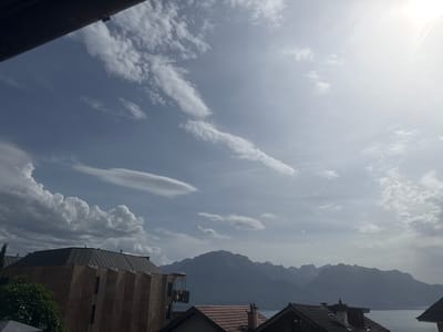 The image captures a scenic view of Montreux, Switzerland, featuring a dramatic sky filled with various cloud formations. In the foreground, the distinctive angular architecture of a modern building is visible, with a green, leafy tree partially obscuring its left side. The building's facade appears to be made of textured stone, and its roof is dark and sloped. Adjacent to this building, a glass-enclosed elevator or capsule is attached to the side, suggesting a unique feature of the structure or its surroundings.
Further into the background, a body of water, likely Lake Geneva, is present, bordered by a distant mountain range. These mountains, shrouded in a blue haze, form a majestic backdrop to the scene. The sky itself is a dominant element, with bright, sunlit areas indicating clear weather interspersed with scattered clouds of different densities and shapes. Some clouds are wispy and stretched across the sky, while others are more voluminous and cumulus-like. The overall impression is one of a partly cloudy day with bright sunlight.